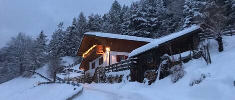 A wooden chalet with a sloped roof, surrounded by snow-covered trees and a wooden fence.