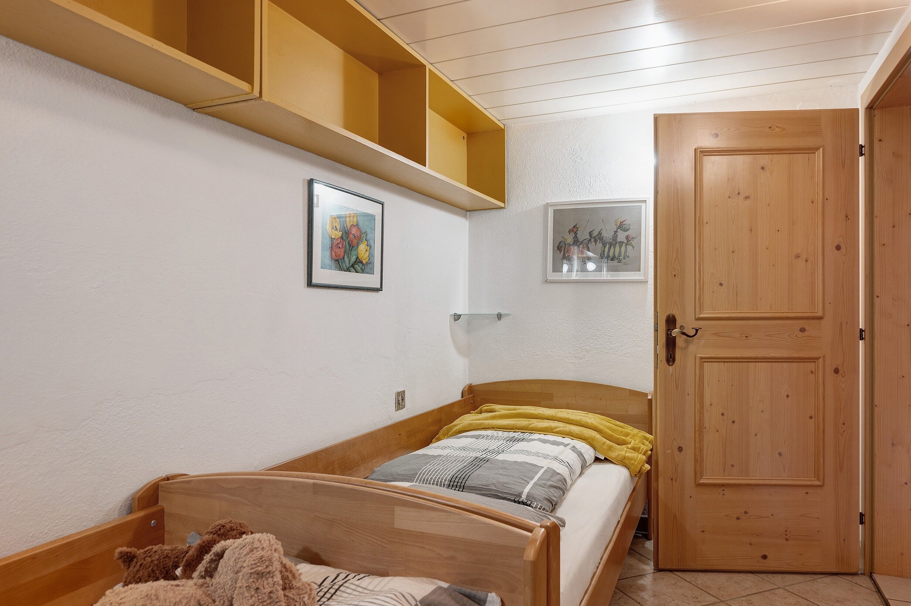 A bedroom with a wooden bed, a shelf above the bed, and a door.