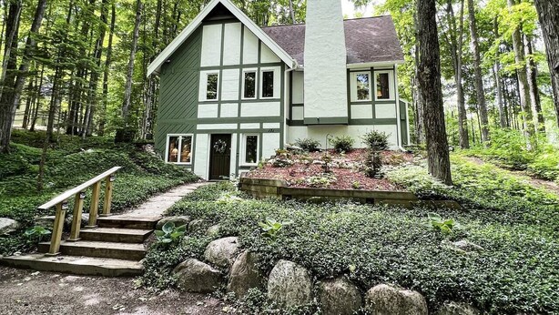 Une maison à deux étages, dotée d’un toit à forte pente, entourée d’une zone boisée.