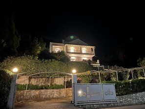 Una casa de dos pisos con balcón, rodeada de follaje y un muro de piedra.