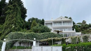 Un bâtiment blanc à deux étages, doté d’un balcon, entouré d’une végétation luxuriante et d’arbres.
