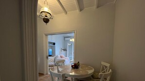 Un coin repas avec une table ronde et quatre chaises, un lustre, et une vue sur une chambre à coucher avec un lit et un téléviseur.