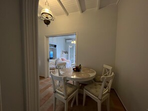 A dining area with a round table and four chairs, a chandelier, and a view into a bedroom with a bed and a TV.
