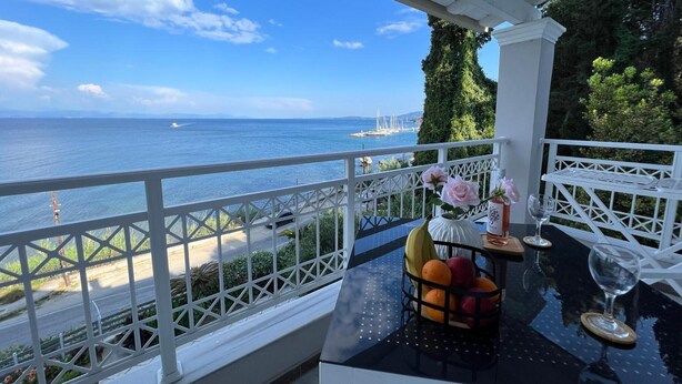 Un balcon avec vue sur la mer, une table avec un panier de fruits et un vase rempli de fleurs.