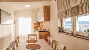 A modern kitchen with wooden cabinets, a window with a view of the sea, and a dining area with a white table and chairs.