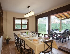 A dining area with a long table set for six, chairs, a window with curtains, and a view of a garden.