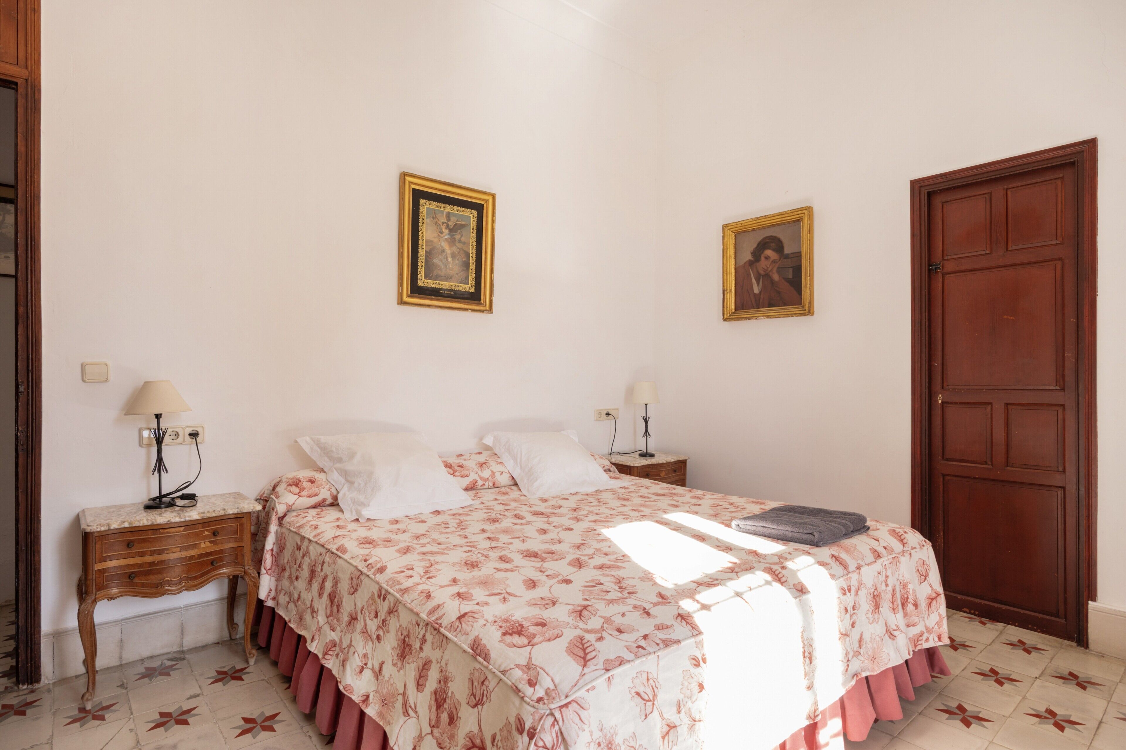 A bedroom with a bed, two bedside tables, a nightstand, a door, and two framed pictures on the wall.