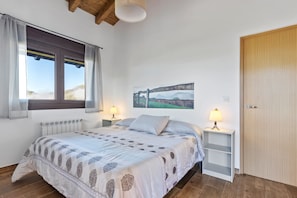 A bedroom with a bed, two bedside tables with lamps, a radiator, a wooden door, and a window with curtains. - Apartment 'Picos' with Mountain Views, Shared Garden, and Wi-Fi (Cangas de onis)