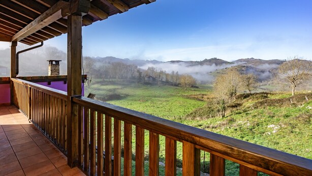 Un balcon en bois offrant une vue sur une vallée verdoyante et des collines enveloppées de brume.