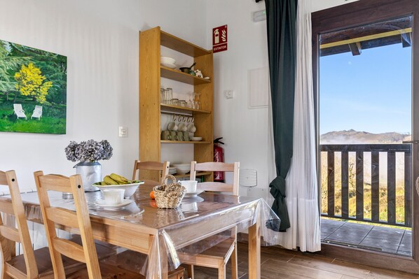 A dining area with a wooden table, chairs, a clear plastic table cover, a wooden shelf with dishes, a fire extinguisher, and a view of the outdoors through a door.