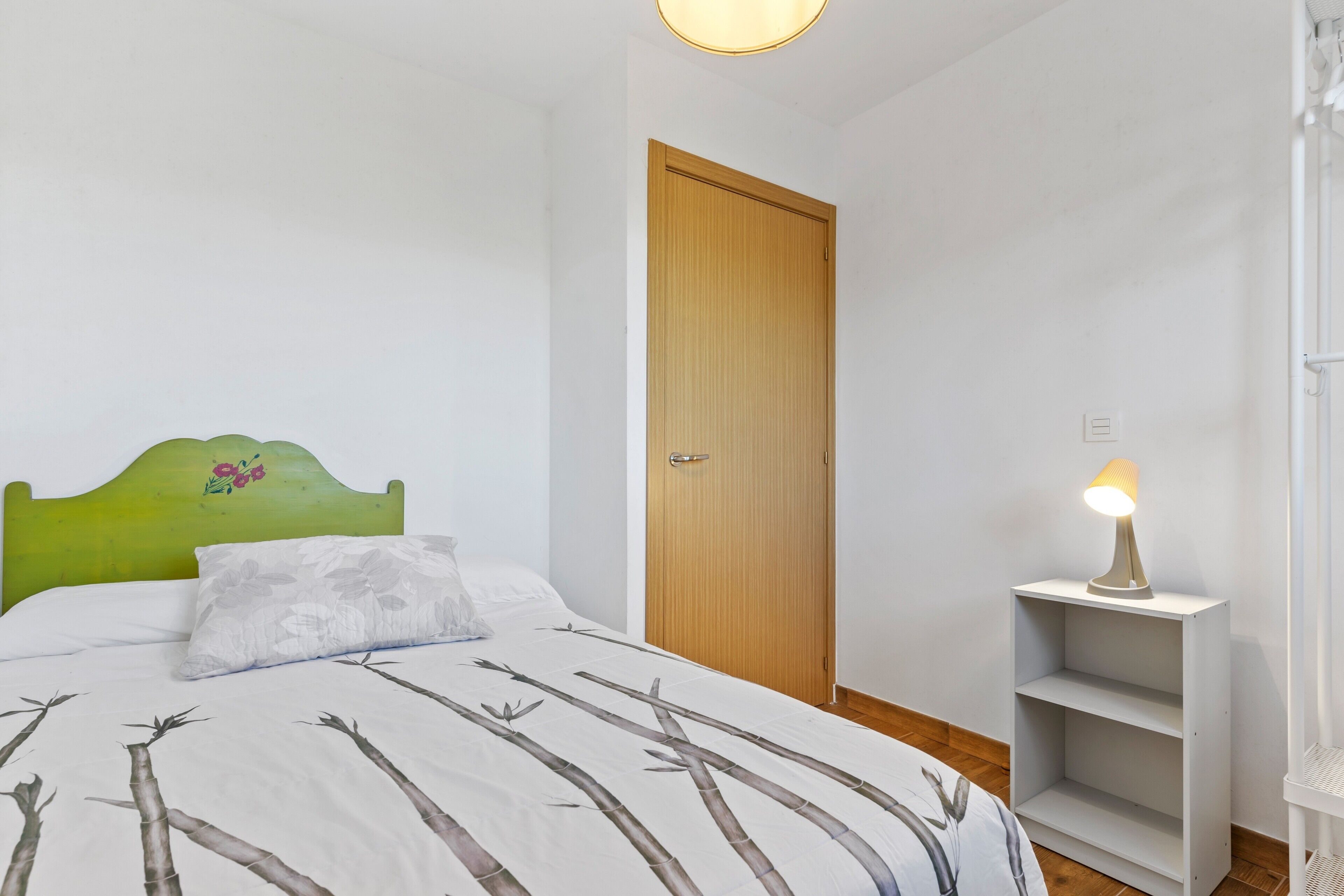 Une chambre à coucher avec un lit, une tête de lit verte, une porte en bois, une table de chevet avec une lampe et une étagère blanche.