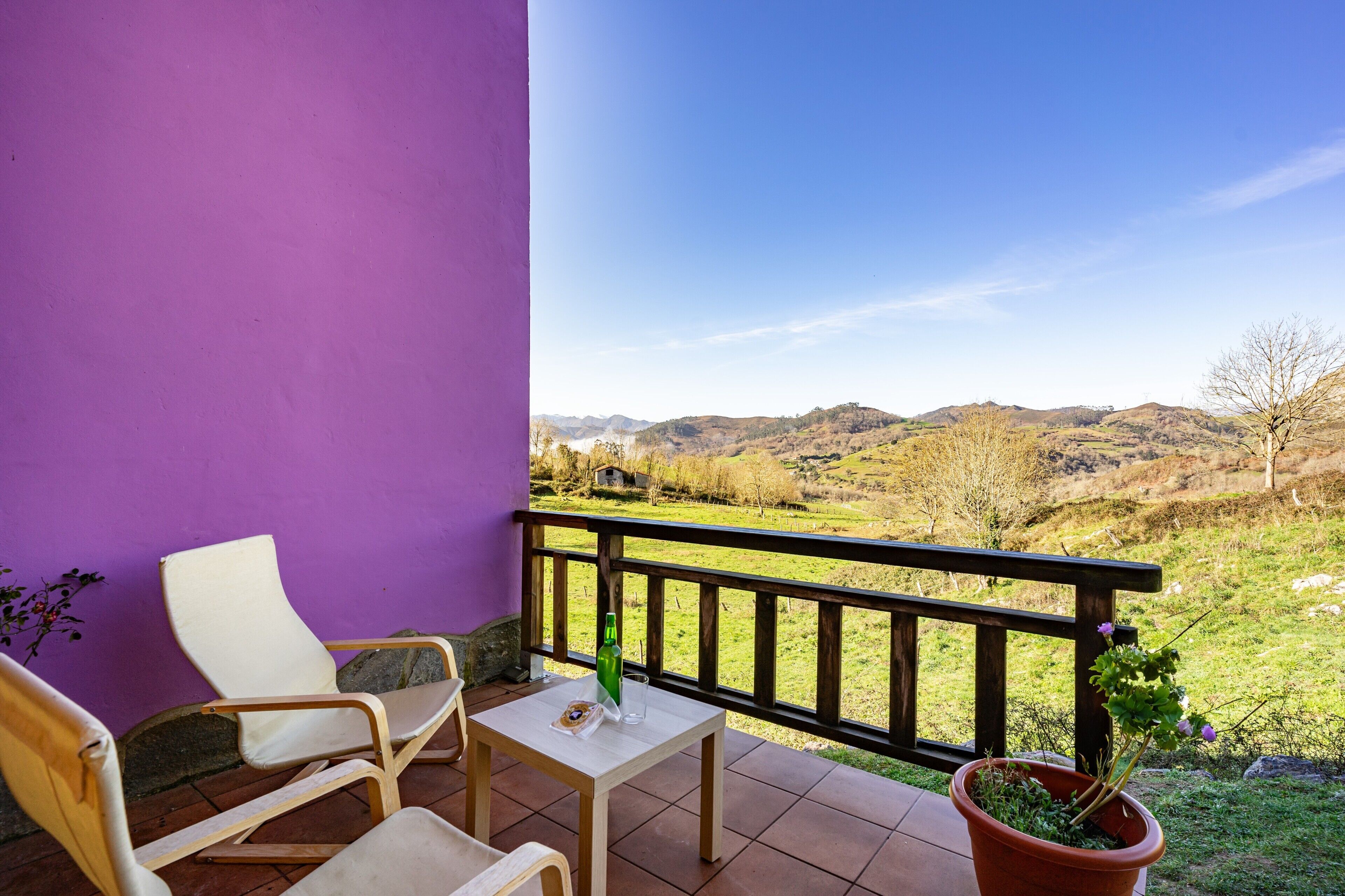 Ein Balkon mit lila Wand, einem Holztisch, zwei weißen Stühlen, einem Blumentopf und einer schönen Aussicht auf Hügel.