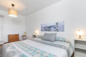 Un dormitorio con una cama, dos mesitas de noche con lámparas, un aparador de madera, una silla y un cuadro colgado en la pared.
