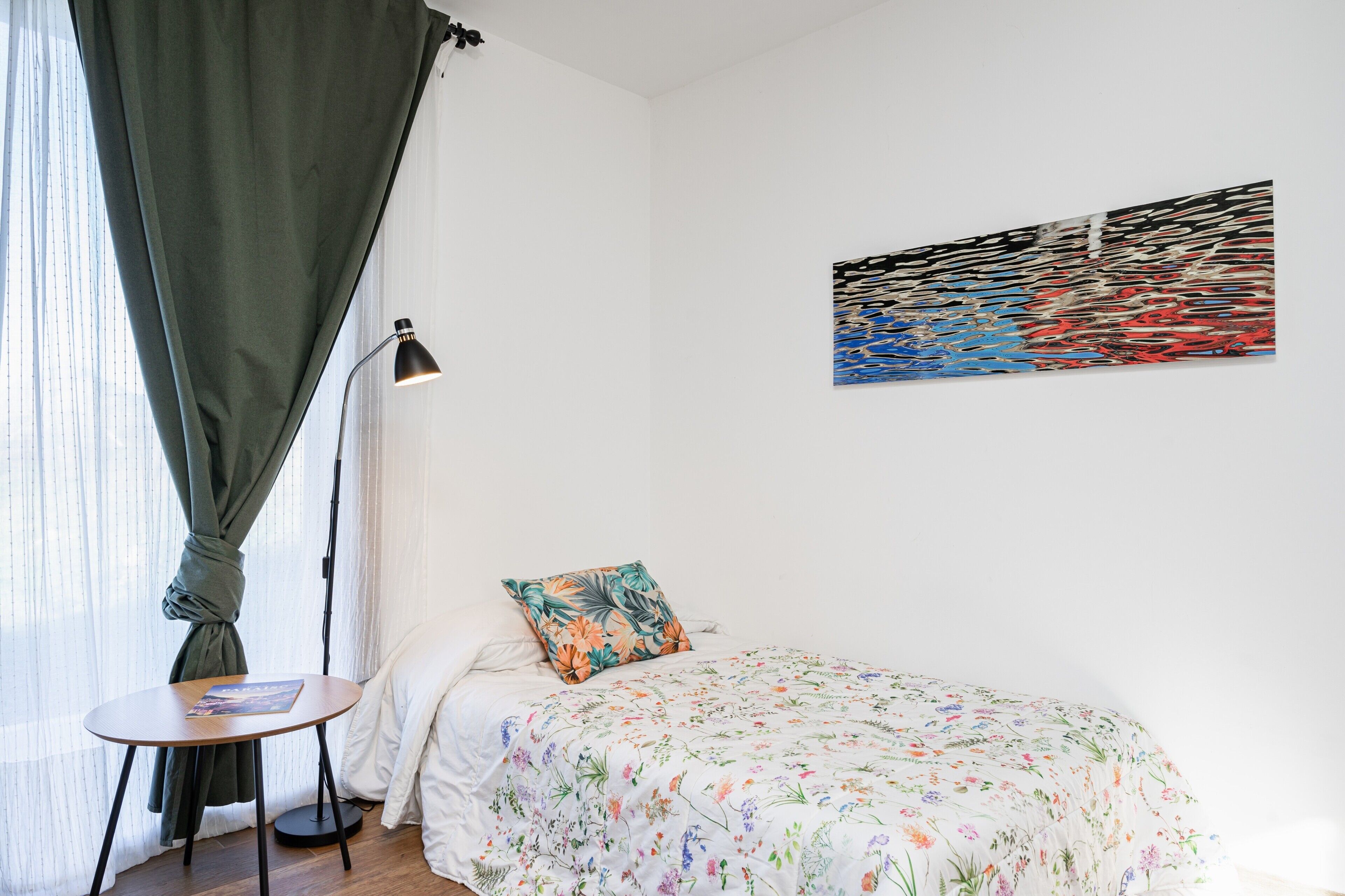 Et soveværelse med en seng, et lille bord, en lampe, et vindue med gardiner og et kunstværk på væggen.