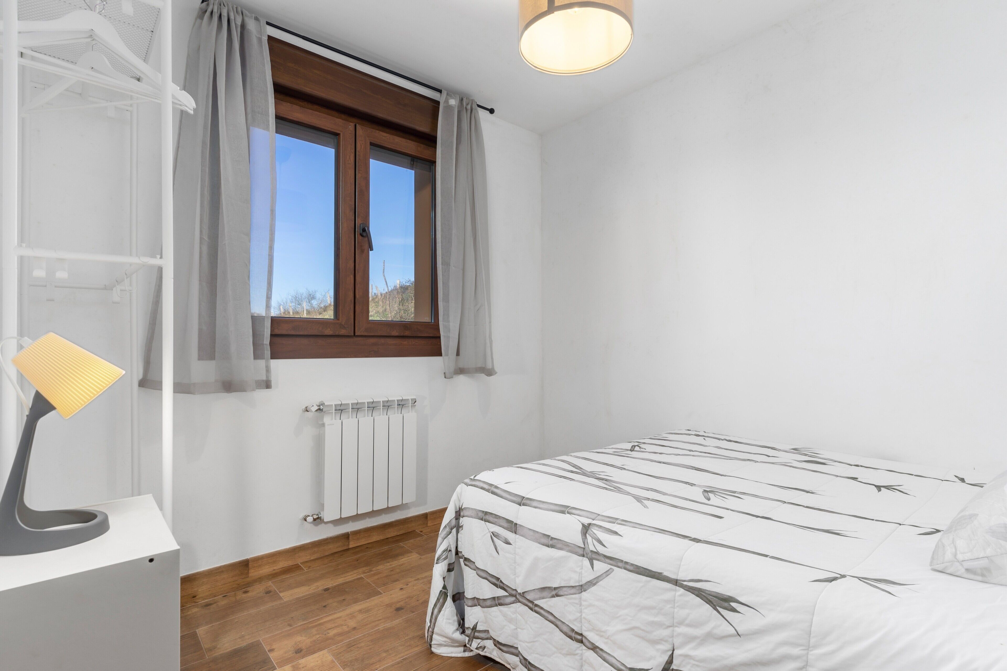 Un dormitorio con una cama, un radiador, una ventana con cortinas y un escritorio con lámpara.