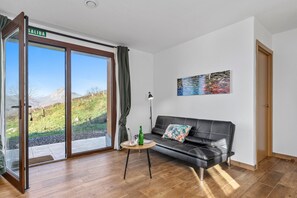 Una habitación moderna con una puerta de vidrio corrediza que da a un balcón, un sofá de cuero negro, una mesita redonda con una botella y una lámpara de pie.