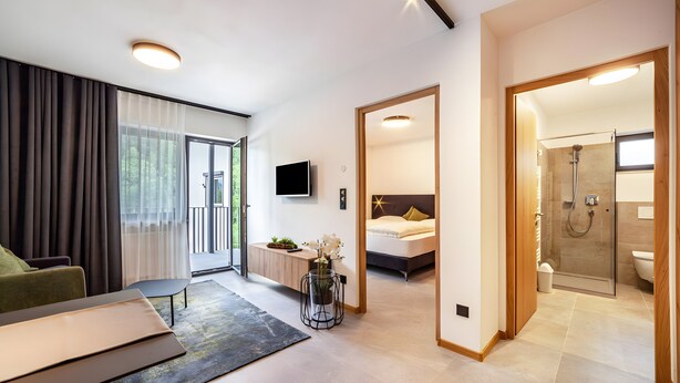 Quarto de hotel moderno com cama, televisão, escrivaninha, sofá, banheiro com chuveiro e varanda com cortinas.