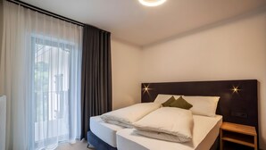Ein modernes Hotelzimmer mit einem großen Bett, einem Nachttisch, Blick durch leichte Vorhänge und einer Deckenleuchte.