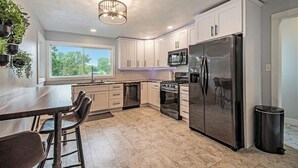 A modern kitchen with white cabinets, a stainless steel refrigerator, a microwave, and a stove. There is a window providing natural light and a ceiling fan.