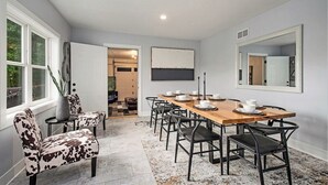 A dining area with a wooden table set for six, surrounded by six chairs. There is a large window on the left, a mirror on the right, and a door leading to another room.