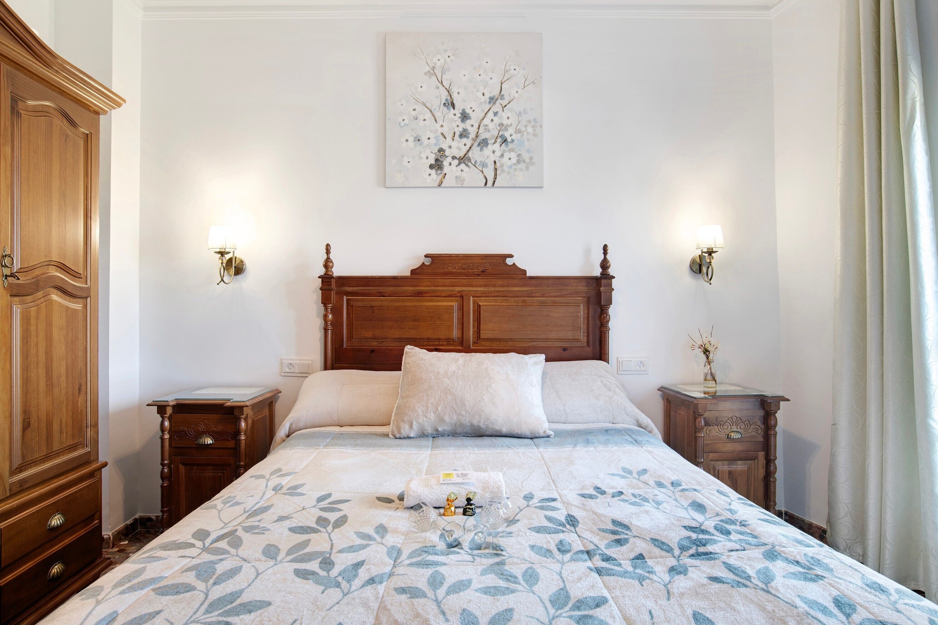 Un dormitorio con cabecera de madera, dos mesitas de noche, una cama con colcha estampada y una lámpara de pared con un florero.