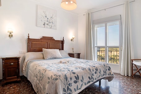 A bedroom with a wooden headboard, a large bed with a floral bedspread, two bedside tables with lamps, a window with a view, and a chair.