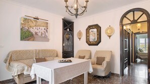 Ein Zimmer mit einer Couch, Sesseln, einem Tisch, einer Chandelüster, einem Wandbild und einer Tür zu einem anderen Raum.