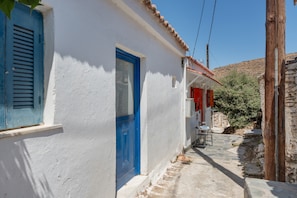 A narrow street with a white building featuring a blue door and shutters, a stone wall, and a small table with chairs outside. - Villa 'Christina 2 Kythnos' with Sea View, Wi-Fi and Air Conditioning (Driopida)