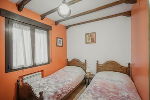 A room with two wooden beds, a radiator, a window with curtains, a ceiling light, and a framed picture on the wall.