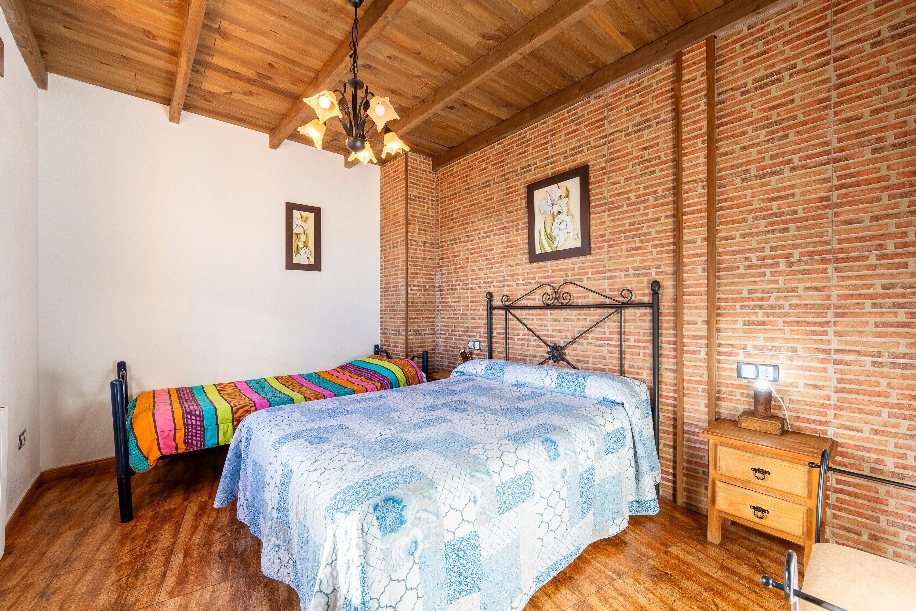 Une chambre avec un plafond en bois, des murs en briques, un lit recouvert d’une couvre-lit à motifs, une table de chevet avec une lampe et un tableau encadré accroché au mur.