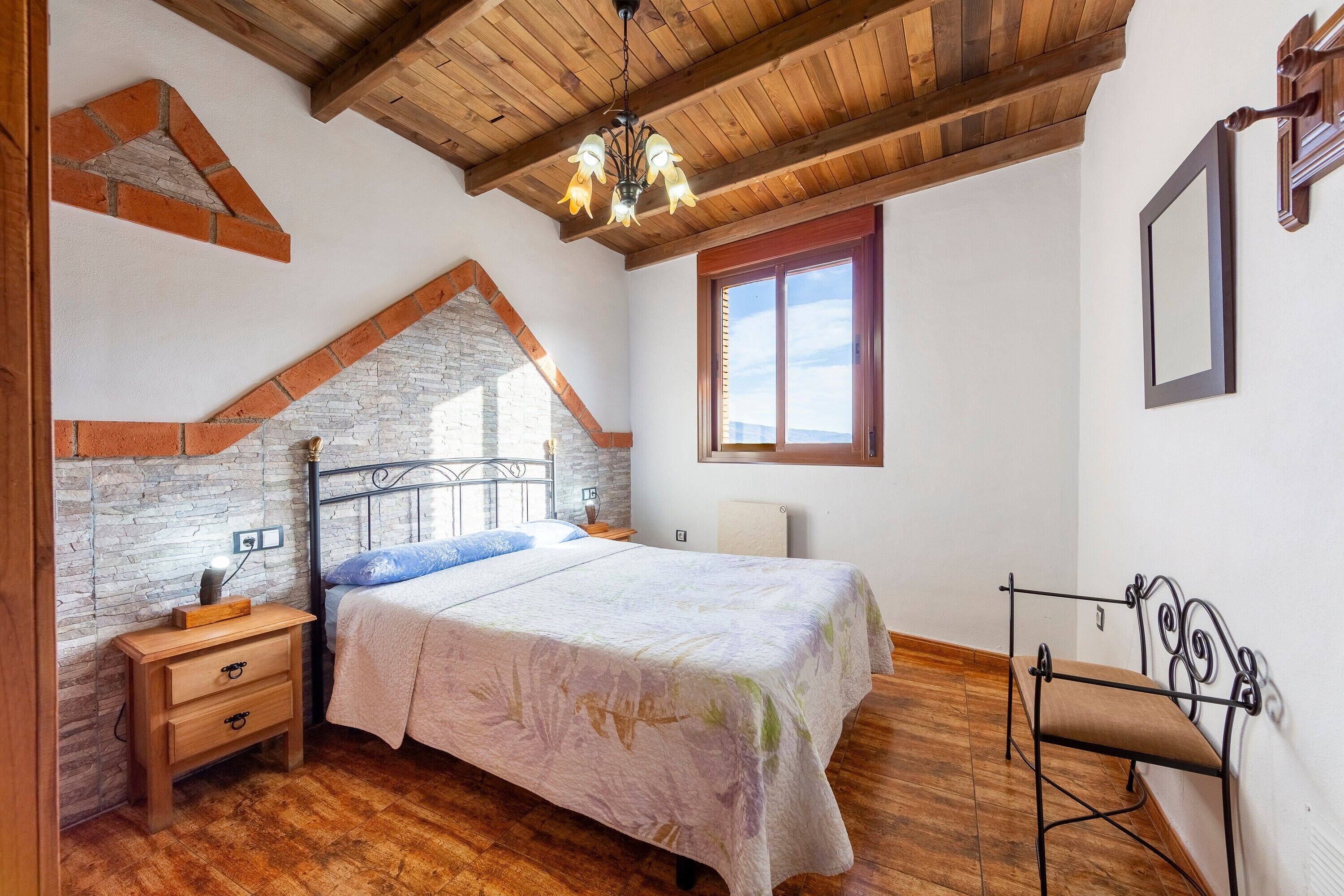 A bedroom with a bed, nightstand, chair, wooden ceiling, brick wall, and a window.
