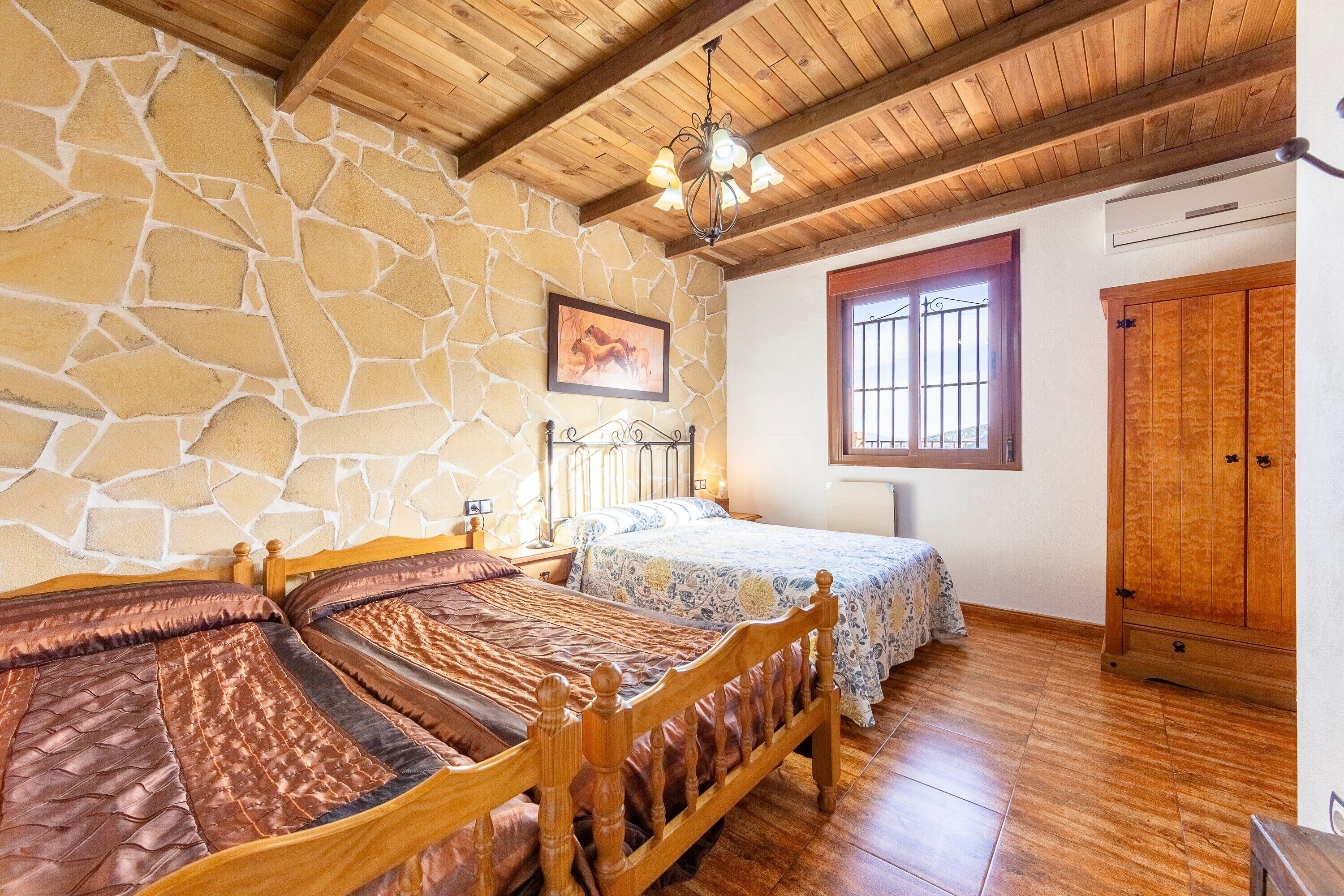A room with a stone wall, wooden ceiling beams, a bed with a headboard, a bedside table, a window with bars, and a wardrobe.
