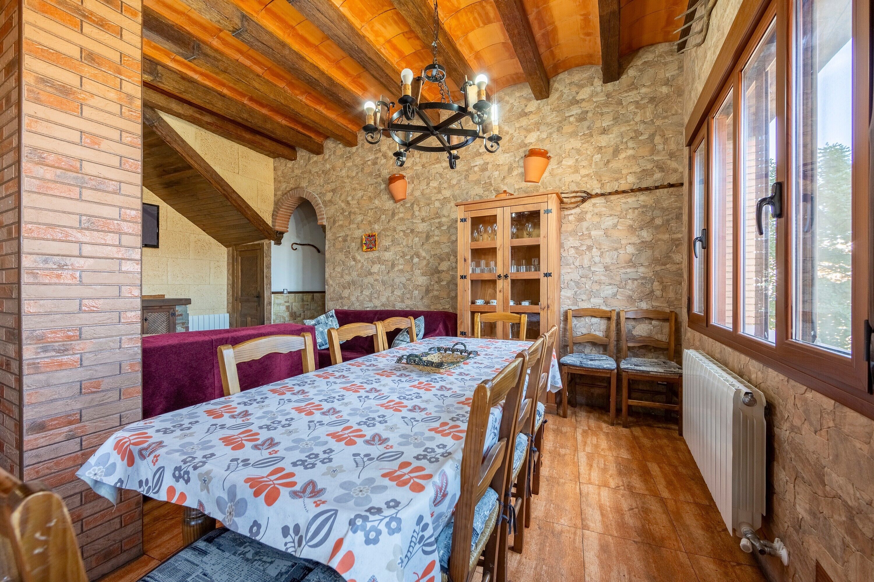 Un comedor con una mesa dispuesta para cuatro personas, sillas de madera, una araña, una pared de piedra y un armario con puertas de vidrio.