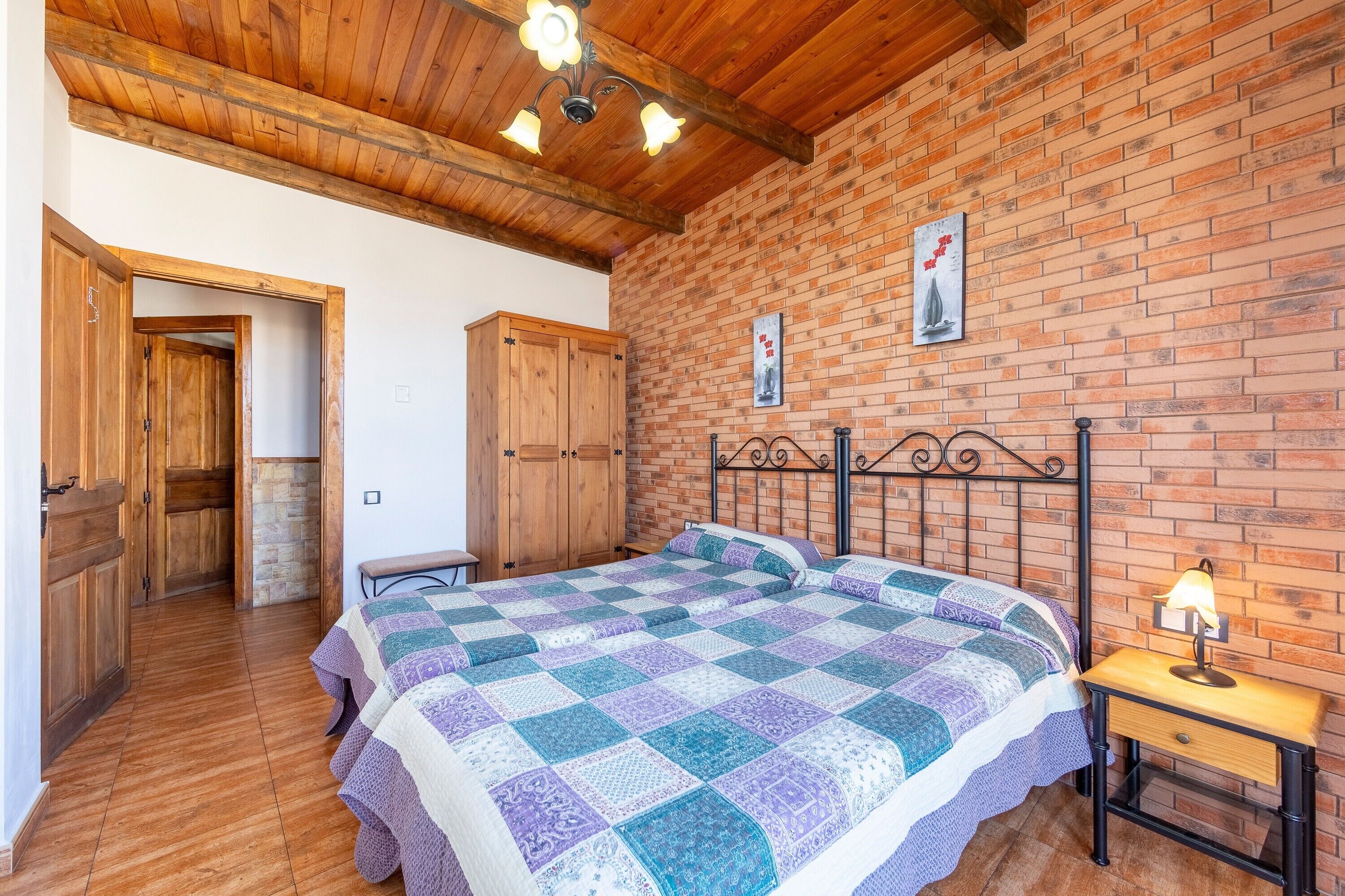 Un dormitorio con techo de madera, pared de ladrillo, una cama con colcha, una mesita de noche y un armario de madera.