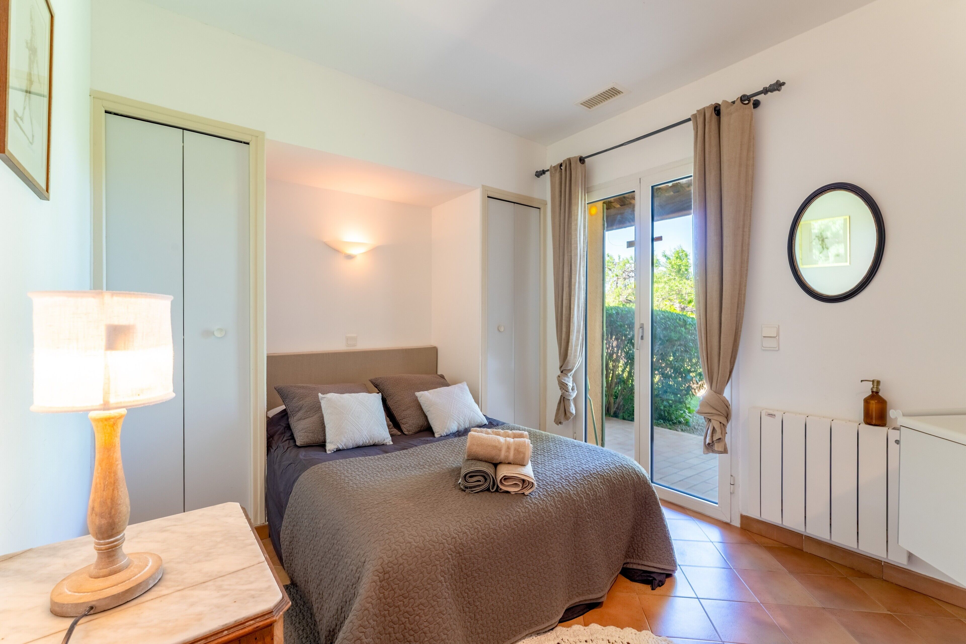 Une chambre à coucher comprenant un lit, une table de chevet, une lampe, un radiateur et une porte-fenêtre coulissante donnant sur l’extérieur.