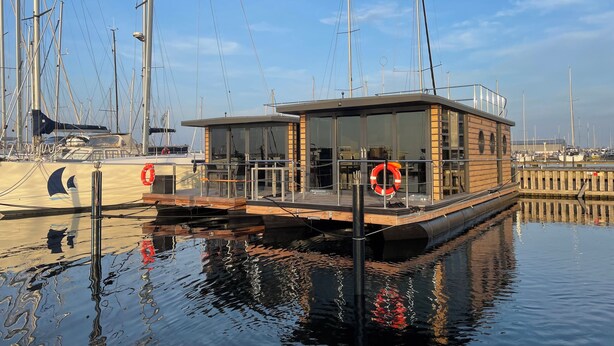 Une structure flottante moderne, dotée d’une façade en verre, amarrée dans une marina où se trouvent de nombreux voiliers.