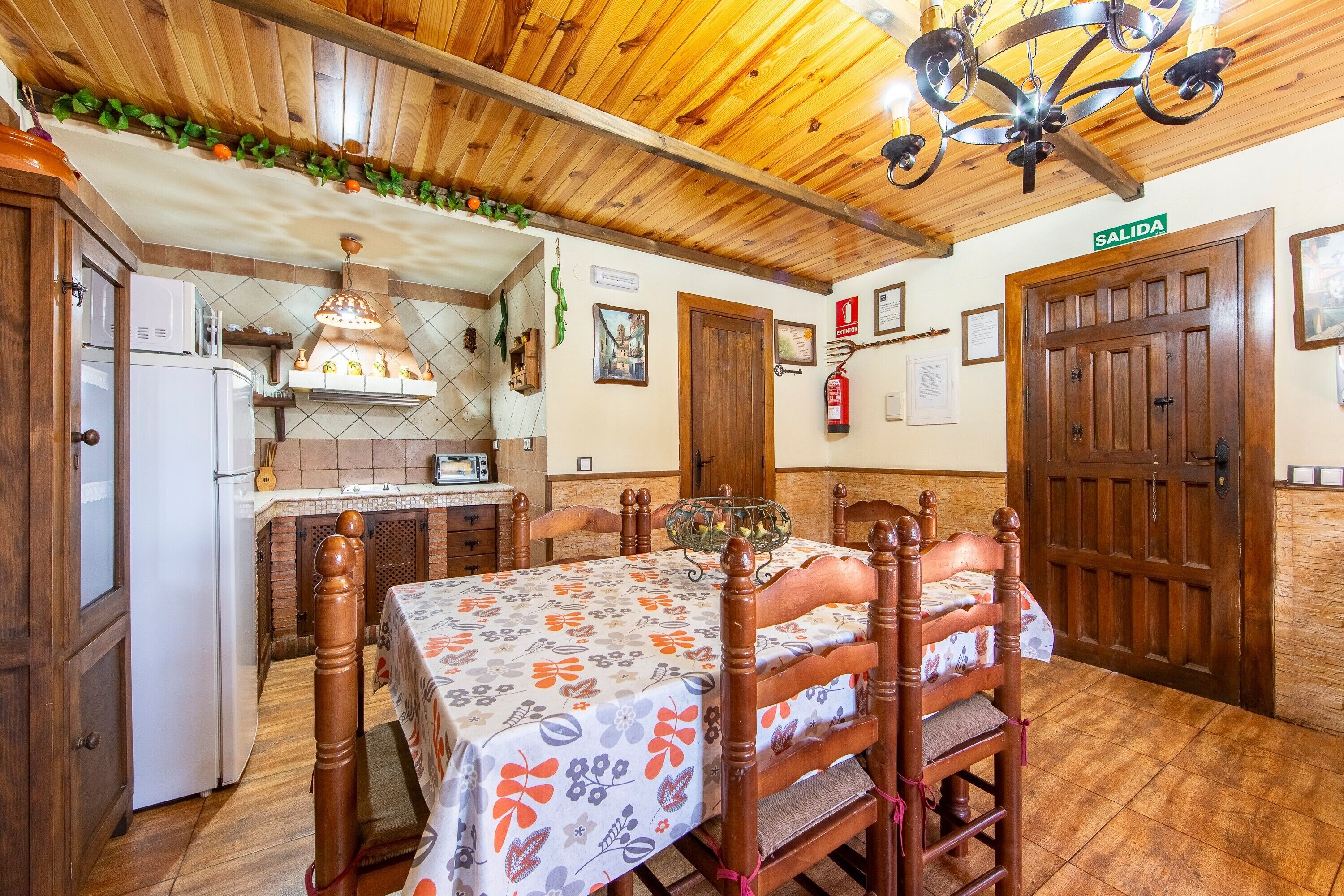 Ein Essbereich mit Holzboden, einem Tisch mit Blumenmuster-Tischdecke, Holzstühlen, einer Pendelleuchte und einer Küche im Hintergrund.
