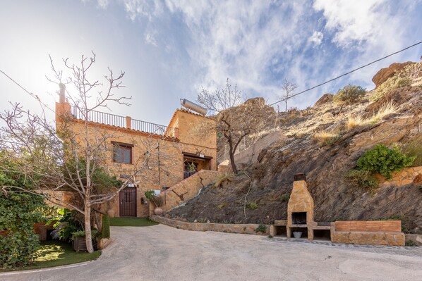 Un edificio de piedra con chimenea, camino empedrado y una ladera rocosa.