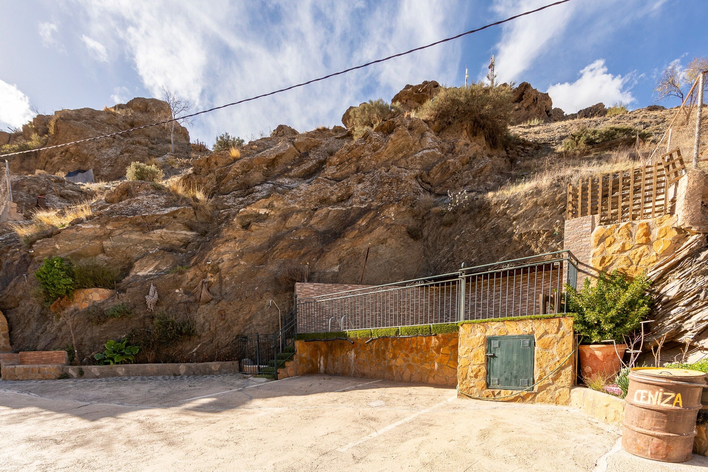 A rocky hillside with a metal staircase leading up, a stone wall with a green door, and a barrel labeled 'CENIZA' in the foreground.