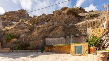 A rocky hillside with a metal staircase leading up, a stone wall with a green door, and a barrel labeled 'CENIZA' in the foreground.