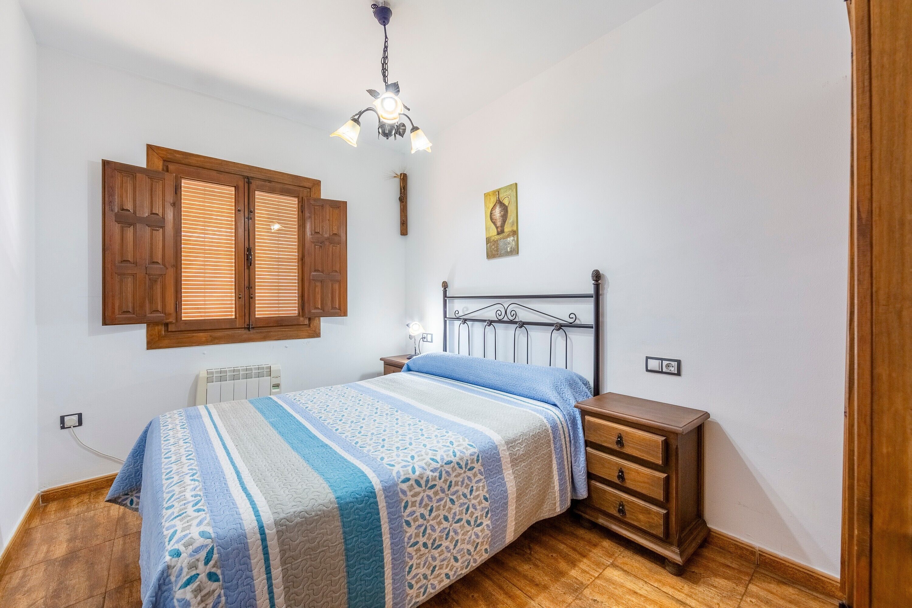 Un dormitorio con una cama, mesitas de noche de madera, un aparador, un radiador, una puerta de madera y una lámpara en el techo.
