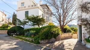 Ein weißes Gebäude mit Balkon, umgeben von Grünpflanzen und einer Steinmauer.