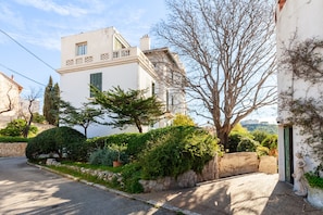 A white building with a balcony, surrounded by greenery and a stone wall.