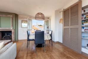 A dining area with a table and chairs, a hanging light fixture, a bookshelf, and a view through a window.