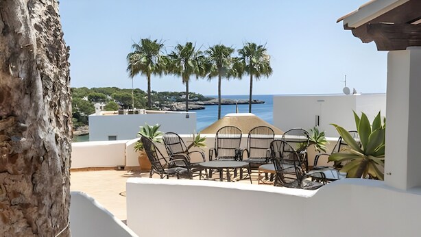 Un área de estar moderna al aire libre con sillas y mesas, una escalera que conduce a un nivel superior, y vista al mar y palmeras al fondo.