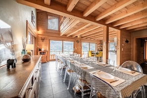 Une longue table à manger, dressée pour un repas, se trouve dans un chalet en bois doté de grandes fenêtres et offrant une vue sur les montagnes.