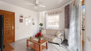 A living room with a sofa, a coffee table with a plant, a ceiling fan, and a wooden door.