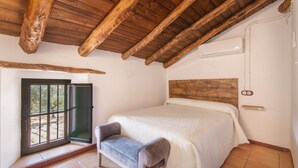Un dormitorio con techo de madera, una cama con cubrecama blanco, un banco y una ventana con vistas a zonas verdes.