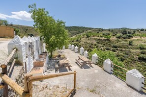 A paved area with picnic tables and a tree, surrounded by white fences and a view of a hilly landscape.