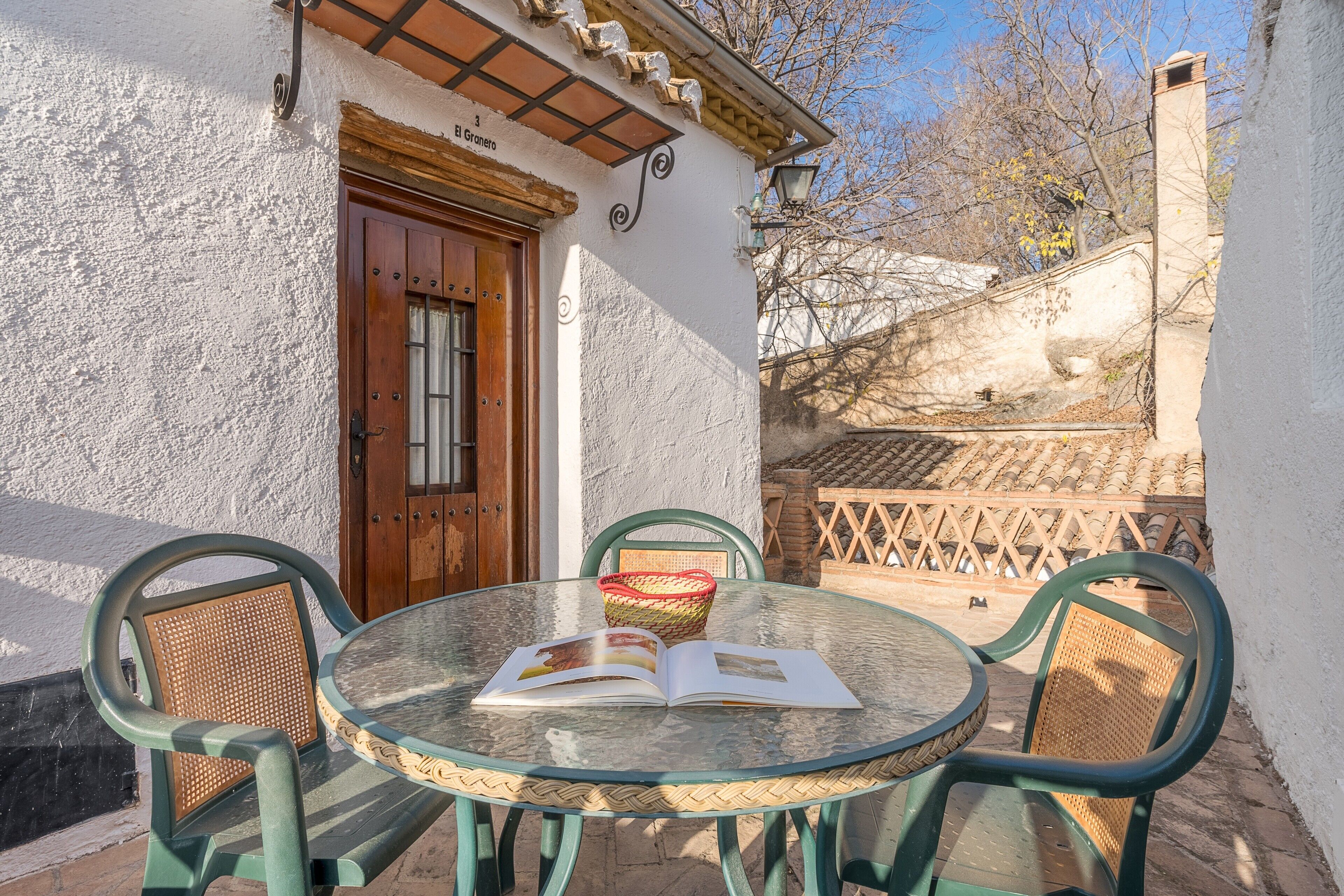 A round glass table with an open book, surrounded by wicker chairs, outside a building with a wooden door and a sign reading 'El Grano'.